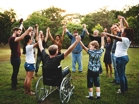An inclusive circle of people are holding hands and raising them above their heads outside. The group includes children, young adults and older adults, as well as a person in a wheelchair.