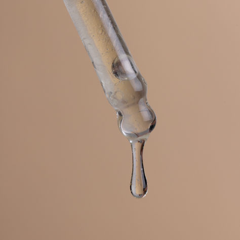 Un primissimo piano macro della punta di un contagocce in vetro da cui pende una goccia densa e trasparente di siero cosmetico o olio, su uno sfondo beige uniforme