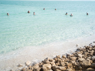 People wading in clear, turquoise water near a rocky shore. Bright, sunny day with a calm and relaxing mood. No text visible.