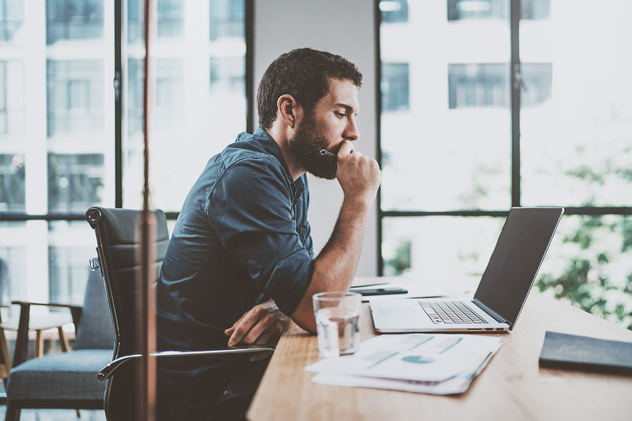 Man working on a laptop at a Work Desk, holding a pen in his hand which is resting on his mouth.
