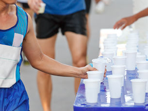 Runner taking a water cup from a table during a race. People in sportswear, cups on blue surface, sunny outdoor setting, energetic mood.