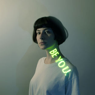 Woman with short dark hair in a white shirt, "BE YOU!" glowing in green on neck. Soft gradient background, reflective mood.