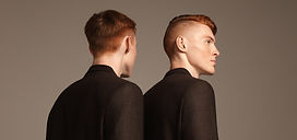 Two male redhead models wearing black suits stand with their backs to each other against a beige background.