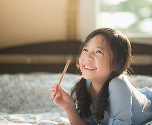 Young girl  lies on her front, holding a pencil, looking upwards and smiling.