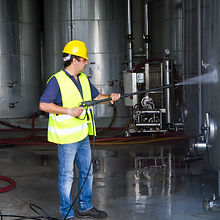 A worker in a yellow hard hat and safety vest uses a pressure washer to clean industrial equipment