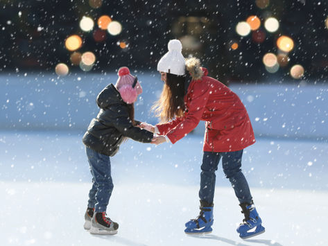 Two people ice skating, one in a red coat and the other in black, holding hands in a snowy rink. Festive lights glow softly in the background.