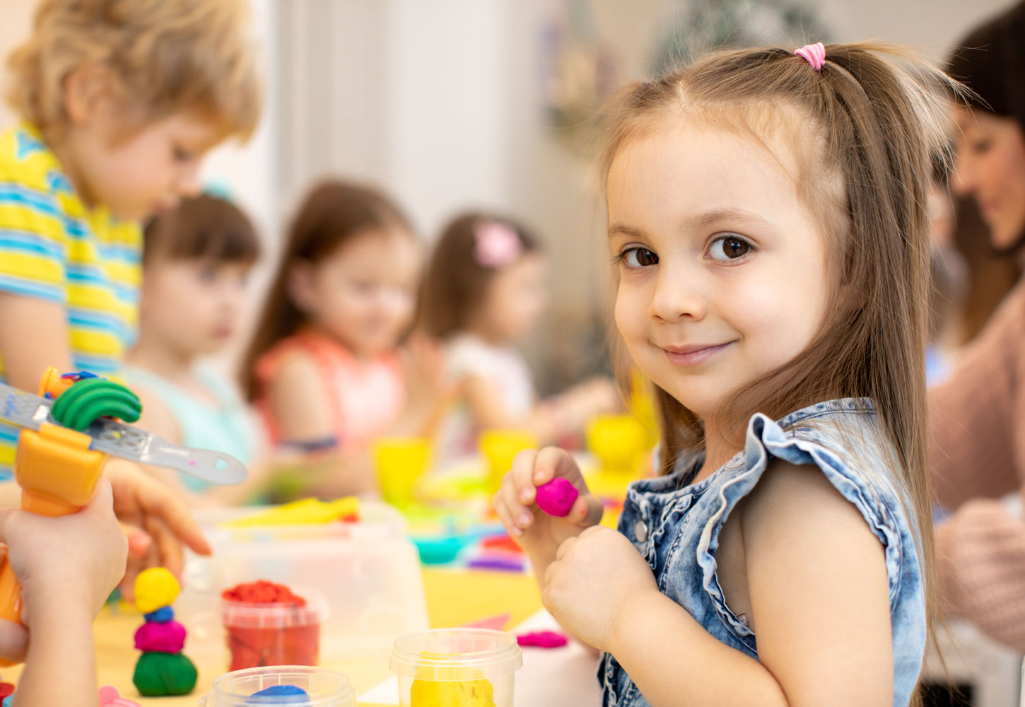 Une petite fille sourit en premier plan pendant un atelier de pâte à modeler, entourée d'autres enfants assis autour d'une table à la crèche Zarafa.