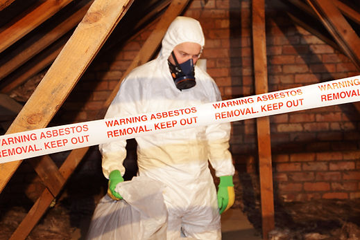 Professional in a white hazmat suit and respirator removing asbestos insulation from pipes in a wooden attic space