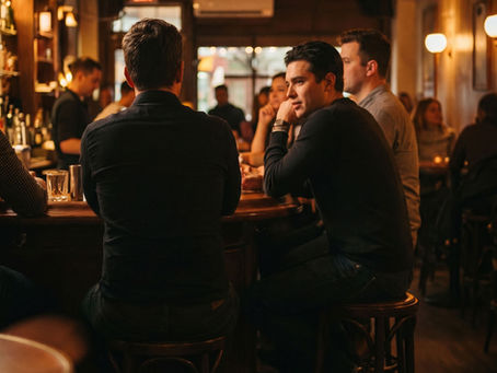 Three men sit on bar stools in a warmly lit pub, chatting amid a blurred crowd; red EXIT sign glows in the background.