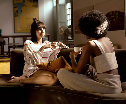 Two women sit facing each other on a couch in a cozy living room, holding mugs and talking. One woman wears a striped long-sleeve top and sits cross-legged, while the other wears a sleeveless outfit and sits with her back partially toward the camera. Soft indoor lighting, pillows, and wall art create a relaxed, intimate setting.