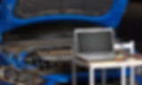 A blue car with an open hood is connected to a laptop displaying graphs on a table