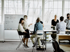 Diverse coworkers discuss a marketing strategy around a table in a bright office, with a whiteboard of notes and a laptop, in a collaborative meeting.