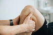 Hands examining a person's knee, suggesting a medical or therapeutic context. The person wears black shorts and a watch. Soft lighting.