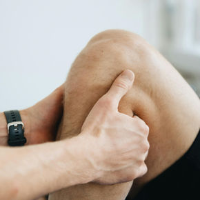 Hands examining a person's knee, suggesting a medical or therapeutic context. The person wears black shorts and a watch. Soft lighting.