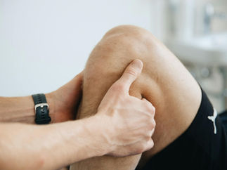 Hands examining a person's knee, suggesting a medical or therapeutic context. The person wears black shorts and a watch. Soft lighting.