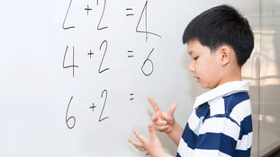 Boy in striped shirt solves math on whiteboard, counting on fingers. Equations include "2+2=4" and "4+2=6". Focused expression.