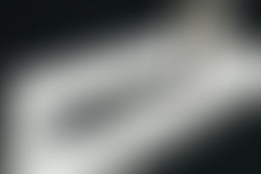 A imagem em preto e branco mostrando a parte inferior do corpo de uma pessoa caminhando em um ambiente iluminado por luz. A pessoa veste uma saia branca com e usa sandálias. A luz projeta uma sombra alongada e bem definida no chão, criando um contraste entre áreas iluminadas e escuras.