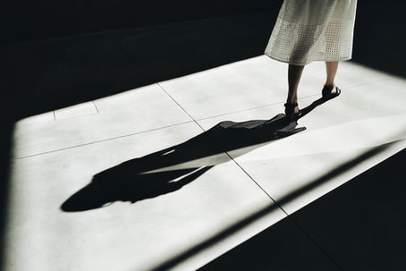 A imagem em preto e branco mostrando a parte inferior do corpo de uma pessoa caminhando em um ambiente iluminado por luz. A pessoa veste uma saia branca com e usa sandálias. A luz projeta uma sombra alongada e bem definida no chão, criando um contraste entre áreas iluminadas e escuras.