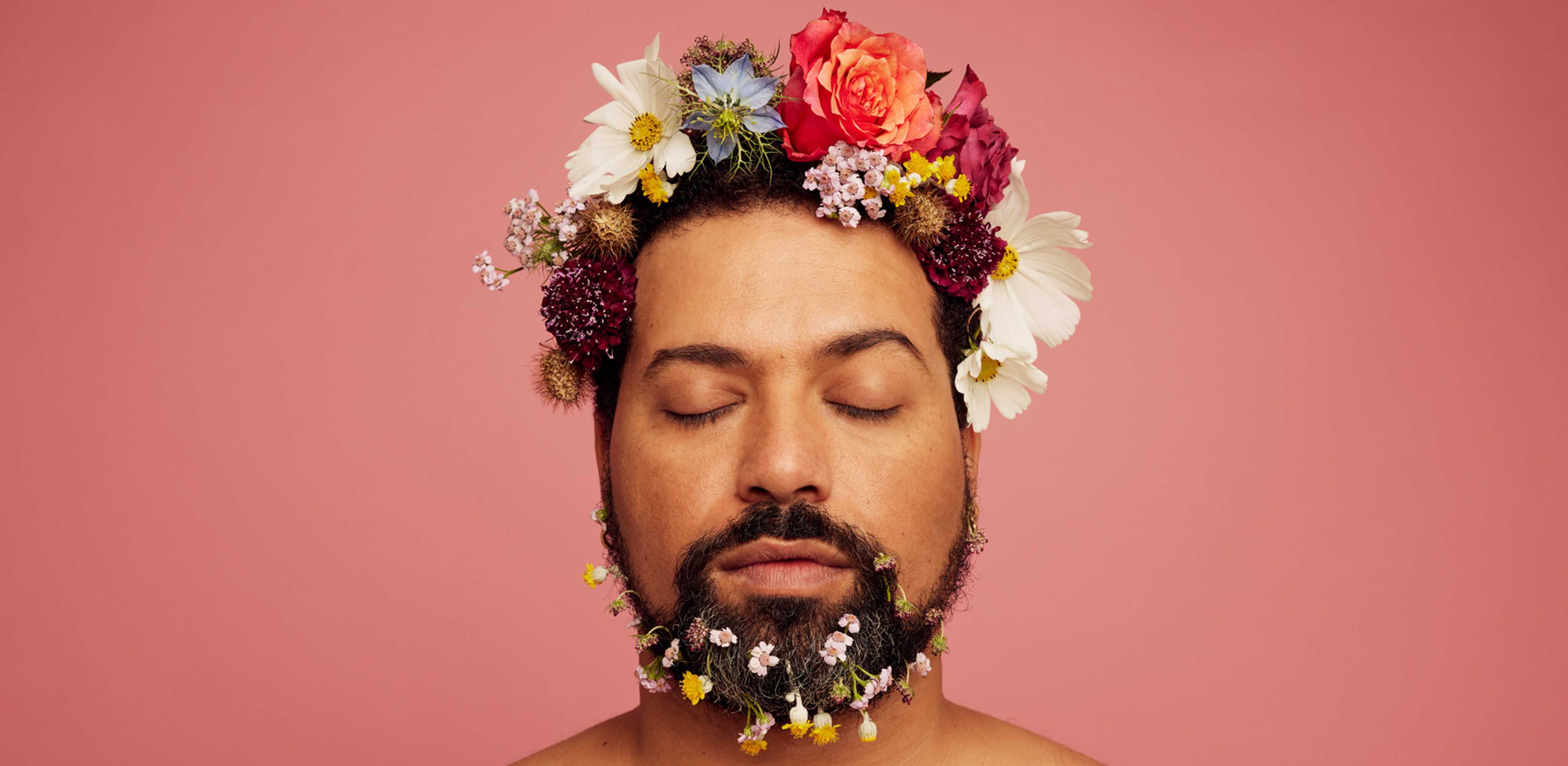 Homme aux yeux fermés, couronne et barbe fleuries, en débardeur blanc sur fond rose, ambiance paisible.