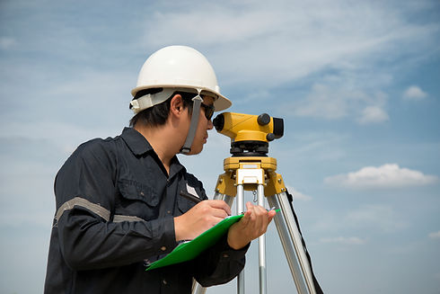 A surveyor wearing a white hard hat uses a theodolite under a blue sky