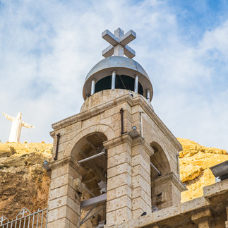 Church on the Mount of Olives overlooking the desert. A traditional stone bell tower featuring a prominent silver cross, with a white statue of Jesus visible in the distance and a barren hillside landscape.