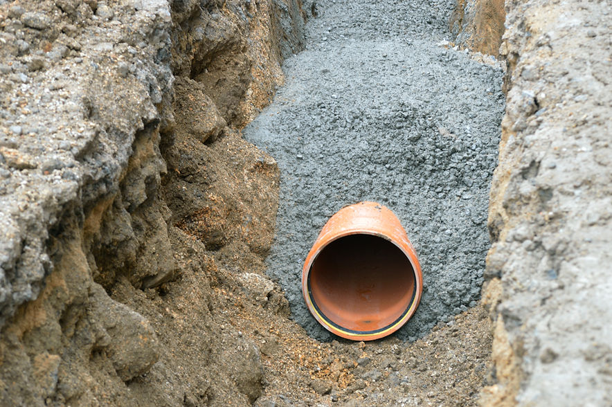 A large clay pipe partially buried in gravel and soil within a narrow, excavated trench