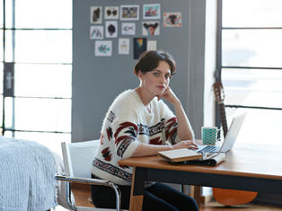 Person in a patterned sweater sits at a table, contemplating, with a laptop, notebook, and mug. Art on walls, bed, and guitar in background.