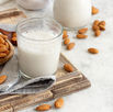 A glass of almond milk on a wooden board surrounded by scattered almonds and a linen cloth. Neutral tones and rustic setting.