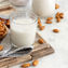 A glass of almond milk on a wooden board surrounded by scattered almonds and a linen cloth. Neutral tones and rustic setting.