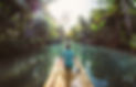 This is an image of a tropical forest there are palms and trees on both sides. The river is calm and fairly narrow. There is a bamboo raft in the middle. There is a man in a hawaiian shirt sitting on the end of the raft in the middle. 