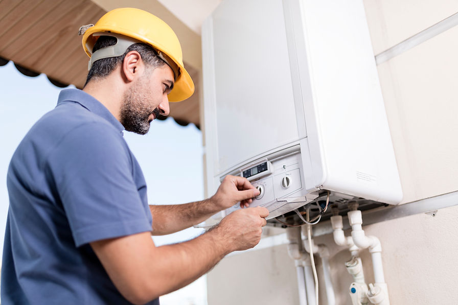 A man in a yellow hard hat and blue shirt inspects and adjusts a white wall-mounted boiler
