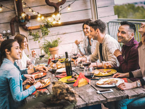 Grupo diverso de quatro pessoas conversando ao redor de uma bancada de madeira texturizada. Sobre a mesa limpa, apenas um prato técnico de crononutrição com salada colorida e proteína, demonstrando o contágio social de hábitos saudáveis.