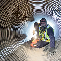 Two people in safety gear crawl through a large, illuminated metal pipe