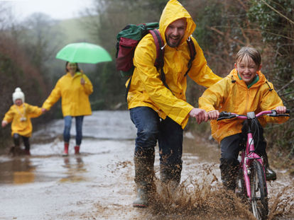 A smiling family cycling together in light rain on a Cornish country lane, wearing raincoats and enjoying the outdoors despite the weather.