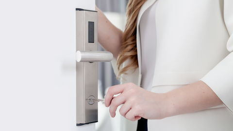 Femme en blanc déverrouille une porte blanche avec une clé, gros plan sur la poignée et la serrure en métal.