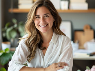 Smiling woman in white shirt at a desk with books and plants on shelves behind. Bright, cozy setting with a welcoming, cheerful mood.