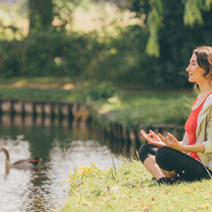 Woman meditating by a pond, sitting cross-legged on grass. Ducks swim nearby. She's wearing a red top and light jacket, serene mood.