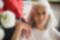 A serene elderly woman with white hair looks at the camera with a person's hand on her shoulder
