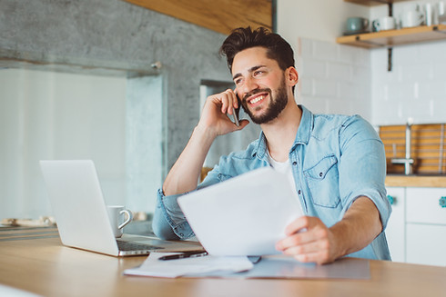 Young man sitting at the table in a kitchen, working on laptop and making phone call. Doin