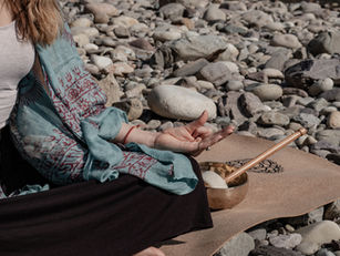 Woman meditating on a rocky beach, wearing a blue patterned shawl. Her hand rests on her knee near a singing bowl and mallet.