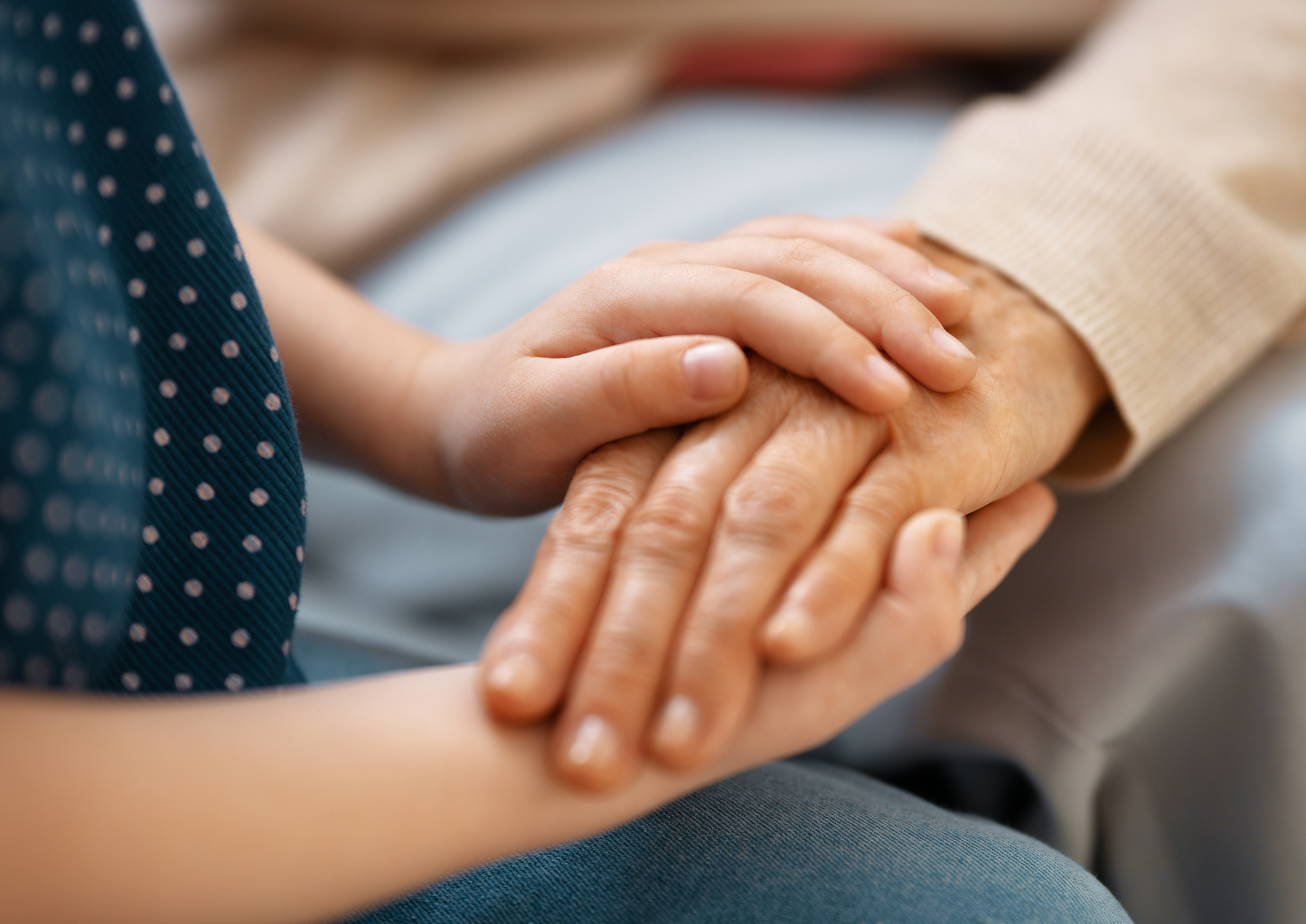 Child’s hand rests on an older person’s hand in a gentle, comforting close-up, with soft blue and beige clothing.