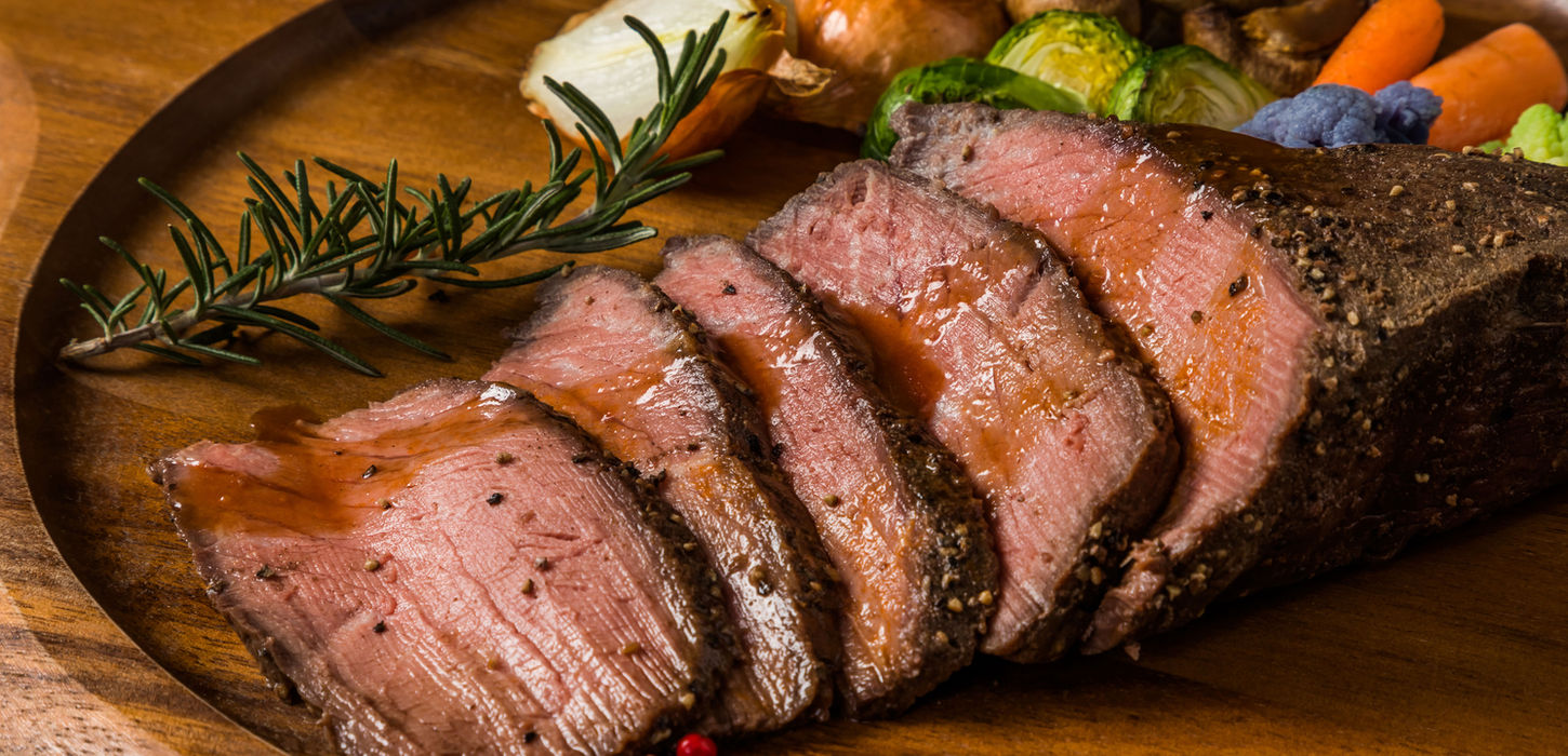 Saftiger, aufgeschnittener Rinderbraten mit Rosmarin und geröstetem Gemüse auf Holzplatte.