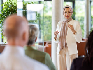 A person wearing beige pants and a beige jacket, with a scarf covering her hair, speaks to a group of people seated in a room with greenery and large windows; a warm, inclusive atmosphere in a workshop meeting.