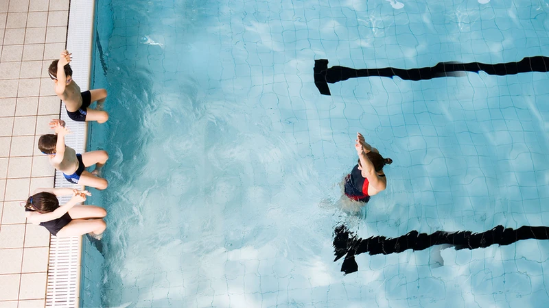 A bird's eye view of a swimming coach in a pool instructing children who are sitting on the edge of the pool about  to dive into the pool. 