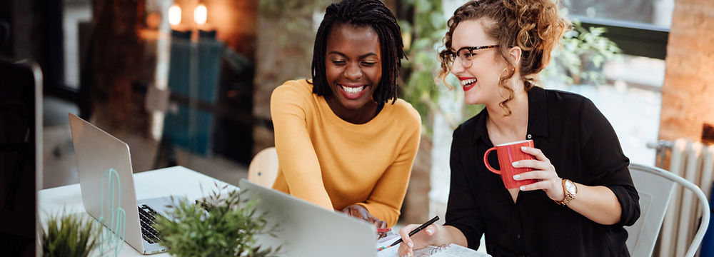 Two women smiling and discussing career development strategies during a coaching session. One is taking notes while the other holds a red coffee cup.