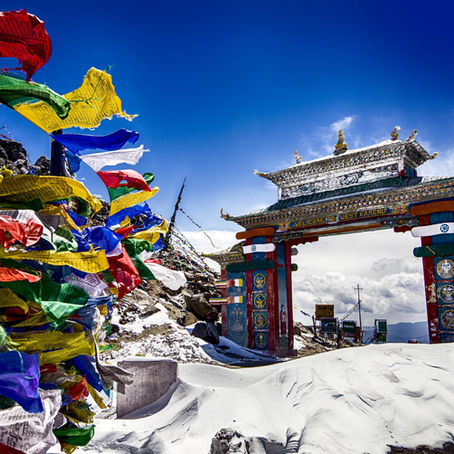 Colorful prayer flags flutter beside a snow-covered mountain pass with an ornate archway under a bright blue sky.