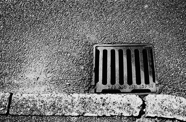 Black and white image of a street drain with vertical bars embedded in rough asphalt