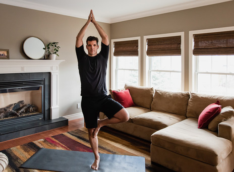 A white man in workout clothes in standing in tree pose with his arms raised to the sky