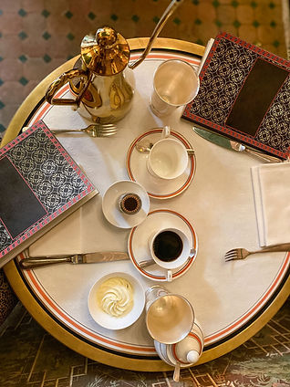 A round table set with a white tablecloth, two ornate menus, a gold coffee pot, a cup of black coffee,