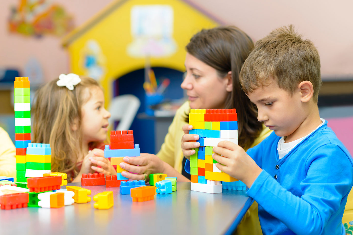 A teacher and two children build colorful block towers at a table in a classroom